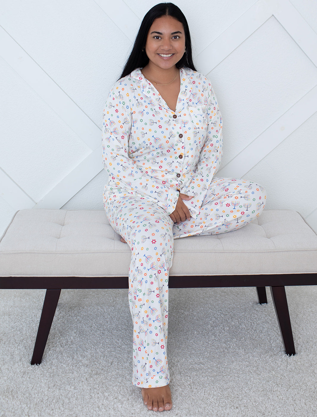 Woman wearing a floral pajama set sitting on a bench against a white geometric wall.