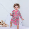 Child in a floral dress standing on a bed with a teddy bear