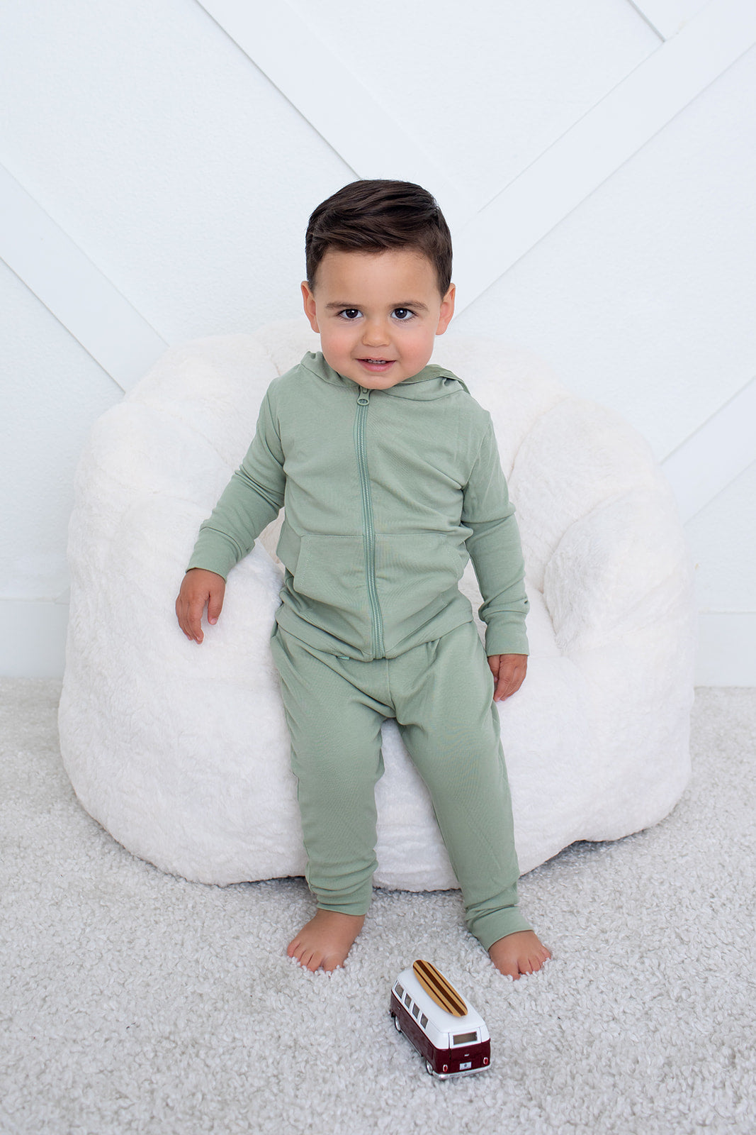 Child wearing a green outfit sitting on a white bean bag chair with a toy train on a light gray floor.