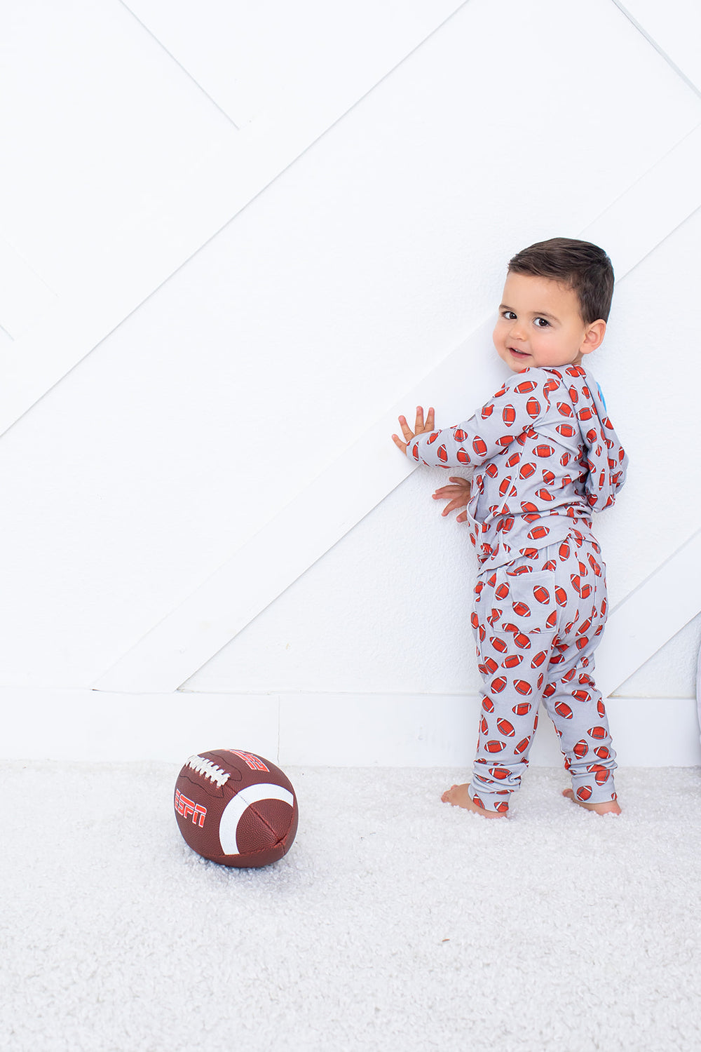 Child in a patterned onesie standing next to a football on a white floor.