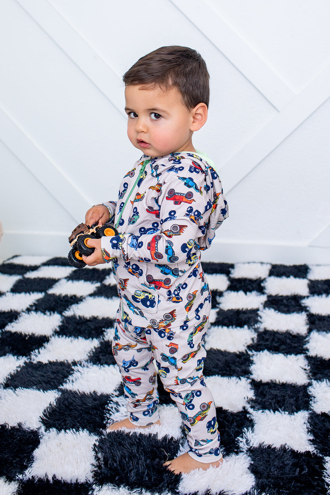 Child wearing a colorful car-patterned onesie on a checkered rug.