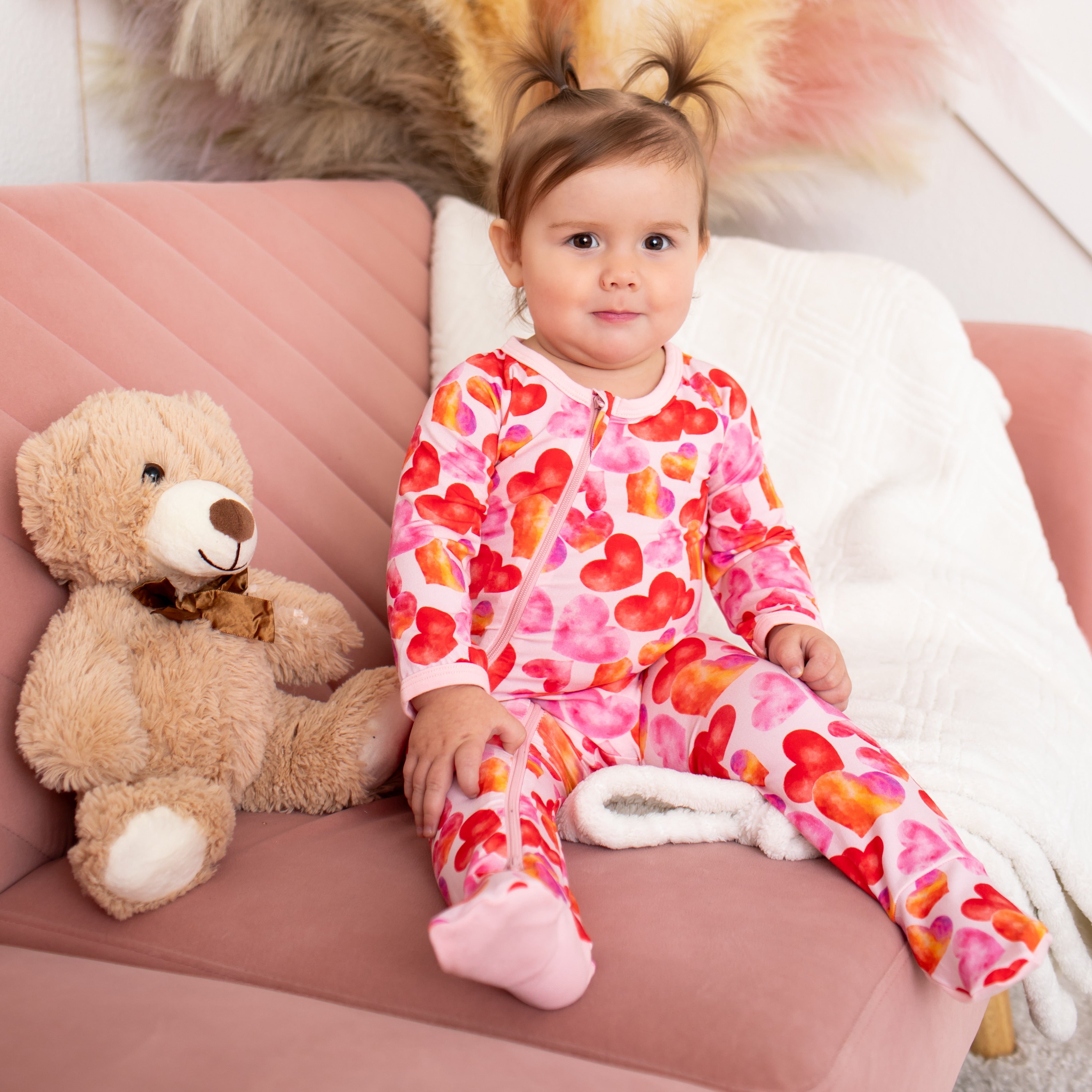 Child in heart-patterned pajamas sitting on a pink couch with a teddy bear and colorful rug.