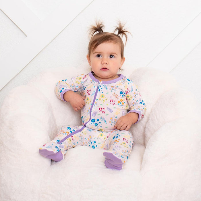 A baby girl with two small pigtails sits in a white plush armchair wearing a long-sleeve footed pajama romper. The pajama has a white base with a colorful floral print and purple trim.