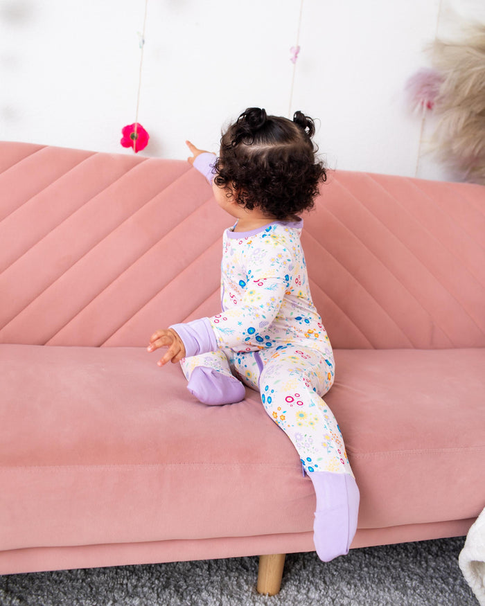 A toddler with curly dark hair sits on a pink sofa wearing a light pastel floral one-piece pajama and lavender socks, pointing at hanging paper flowers on a white wall.