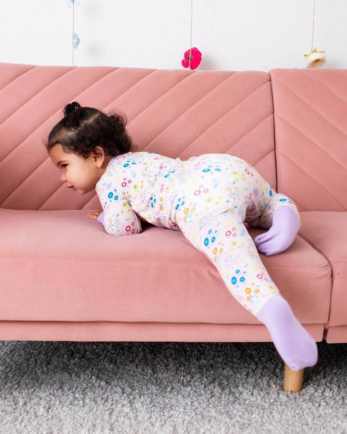 A young child wearing a floral print one-piece pajama and light purple socks is crawling on a dusty pink quilted sofa in a brightly lit room.