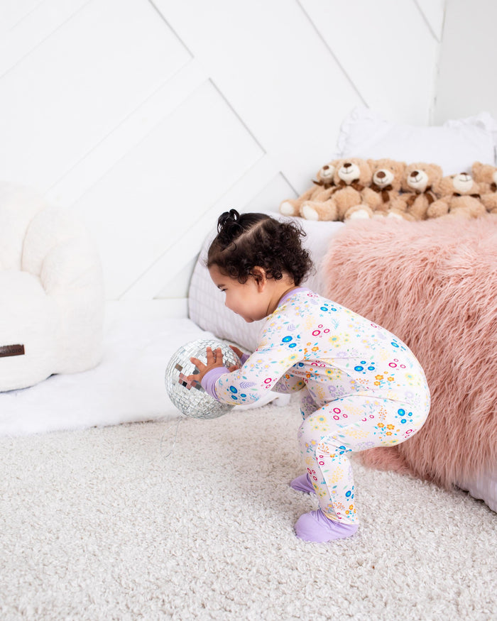A toddler girl wearing a white long-sleeve pajama set with a colorful floral pattern crouches on a light gray carpet in a bright bedroom, holding a plush toy near a pink faux-fur blanket and teddy bears.