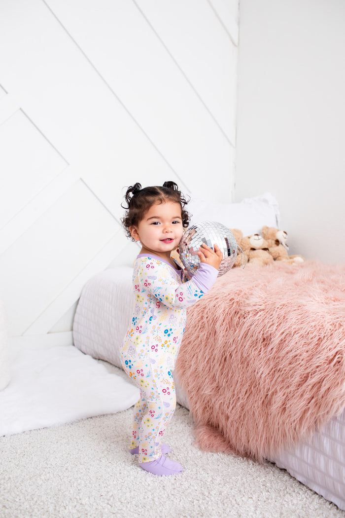 A toddler girl stands in a bedroom wearing a light pastel floral one-piece pajama and holding a silver toy. The room features a white paneled wall, a fluffy pink blanket, and a white quilted bed.