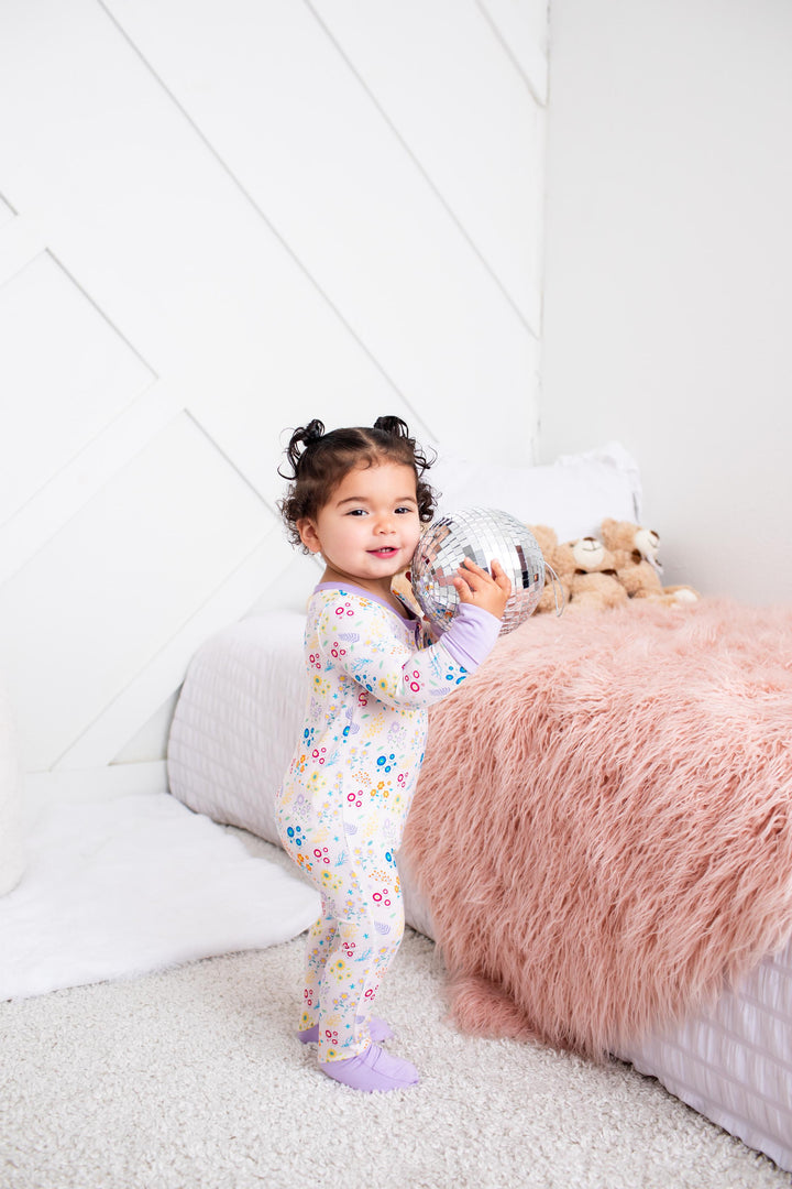 A toddler girl stands in a bedroom wearing a light pastel floral one-piece pajama and holding a silver toy. The room features a white paneled wall, a fluffy pink blanket, and a white quilted bed.