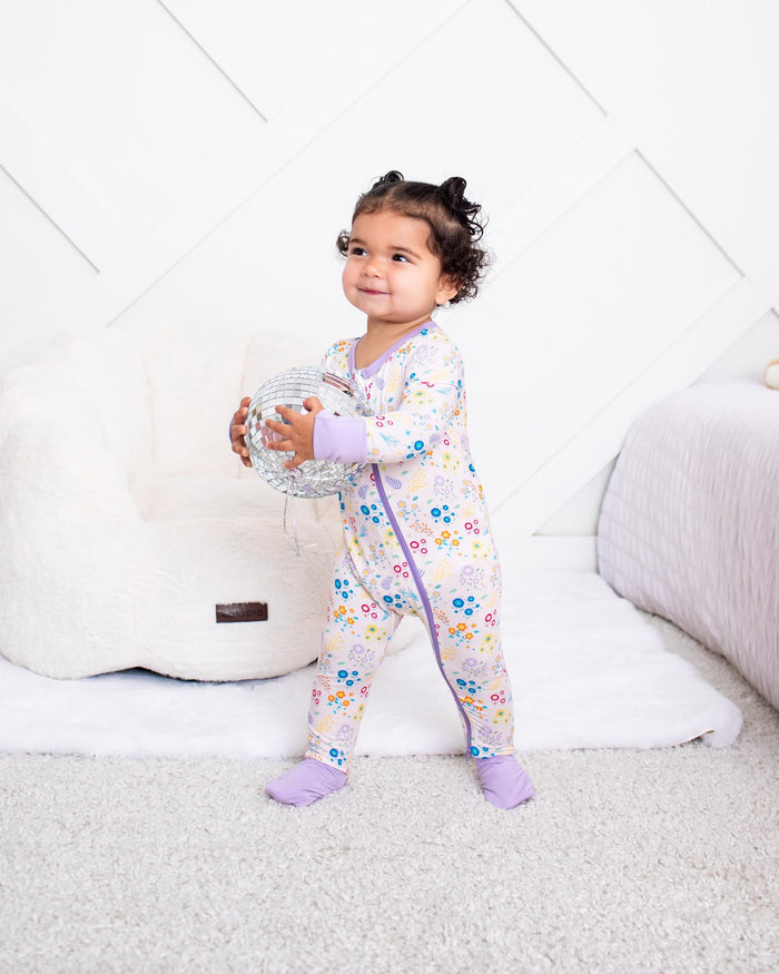 A toddler girl stands in a brightly lit nursery wearing a light pink floral long-sleeve pajama romper and purple socks, holding a clear ball. The background features a white geometric paneled wall, a white bean bag chair, and a bed with a pink blanket.