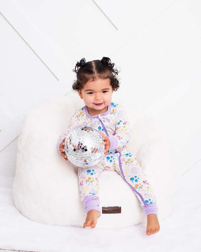 A toddler girl with her hair in two buns sits on a white beanbag chair, wearing a light purple and white floral pajama set and holding a silver disco ball.