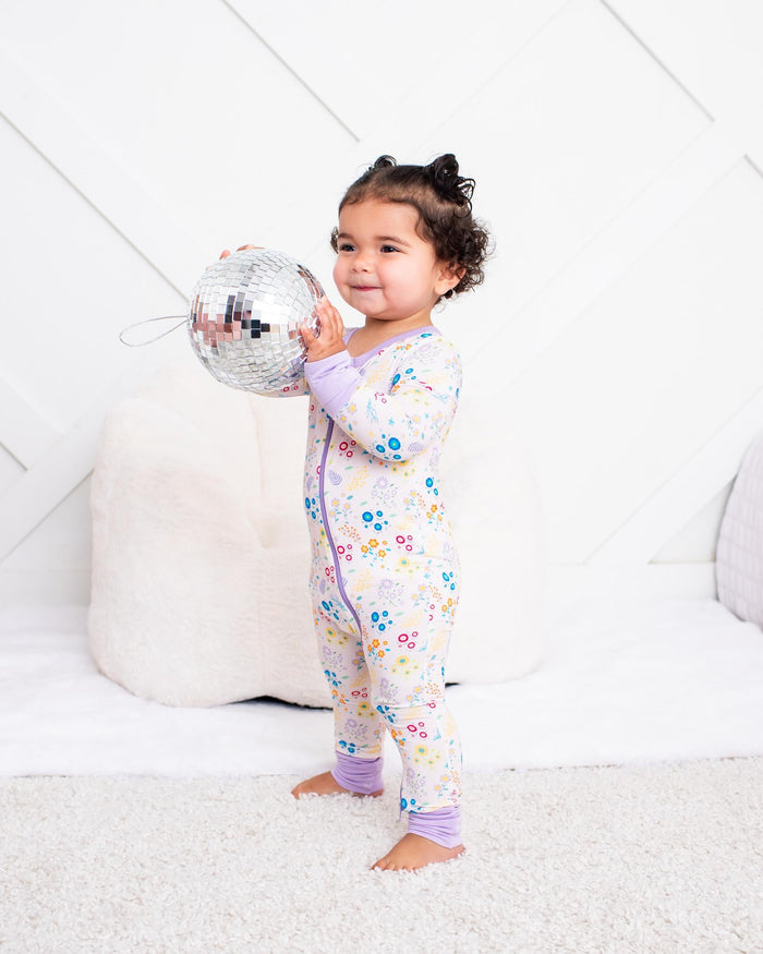 A toddler girl stands barefoot in a bright, modern bedroom setting, holding a silver disco ball. She is wearing a light purple and white floral one-piece pajama top and pants set.