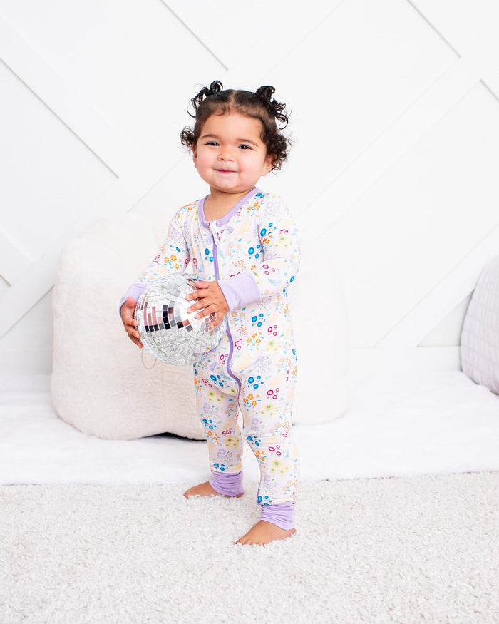 A toddler girl stands barefoot in a brightly lit nursery wearing a light lavender and white floral long-sleeve pajama set. She is holding a small toy, with a white floor mat, pillows, and a bed with a pink blanket in the background.