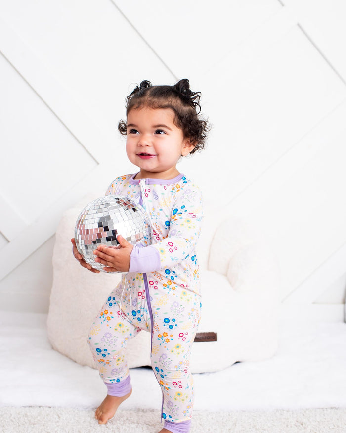 A toddler girl stands barefoot on a white rug in a bright room, wearing a light pink floral pajama set and holding a silver disco ball.