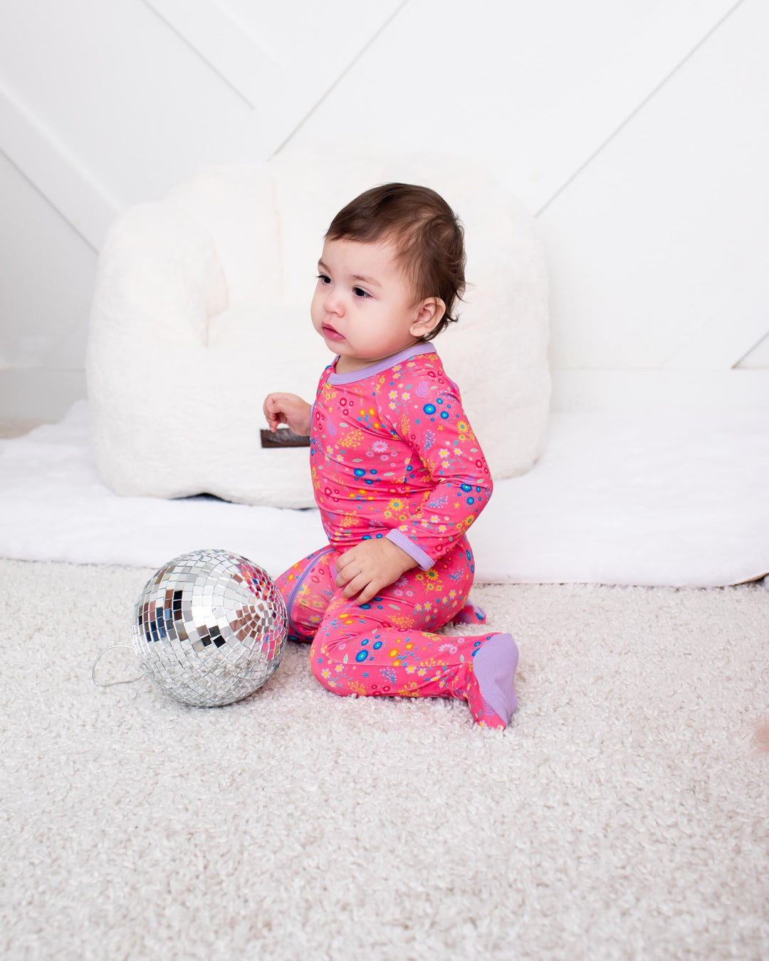 A baby sitting on a white carpet in front of a white paneled wall, wearing a pink long-sleeve pajama set with a colorful pattern. A silver disco ball toy is on the floor next to the baby.