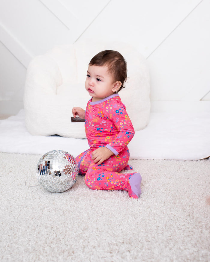 A baby sitting on a white carpet in front of a white paneled wall, wearing a pink long-sleeve pajama set with a colorful pattern. A silver disco ball toy is on the floor next to the baby.