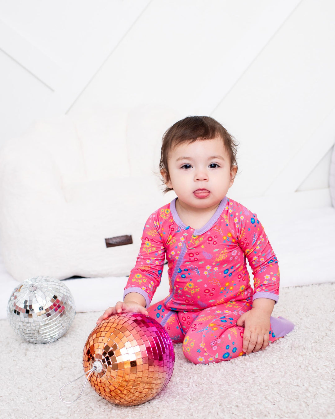 A toddler girl wearing a pink floral long-sleeve pajama set sits on a white carpeted floor, holding a shiny orange and pink disco ball. A smaller silver disco ball is on the left, with a white padded play couch and a pink fuzzy blanket in the background.