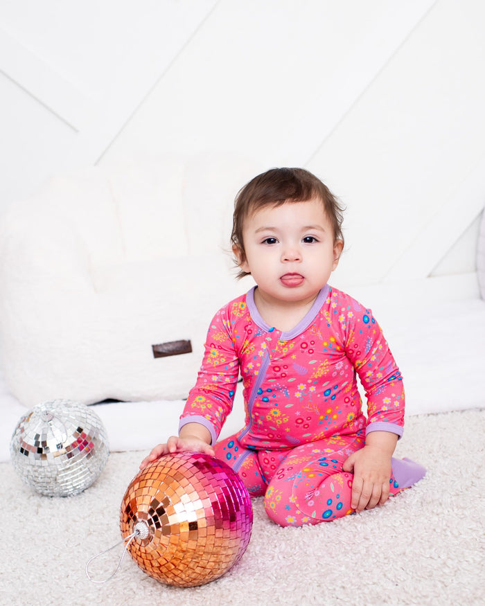 A toddler girl wearing a pink floral long-sleeve pajama set sits on a white carpeted floor, holding a shiny orange and pink disco ball. A smaller silver disco ball is on the left, with a white padded play couch and a pink fuzzy blanket in the background.