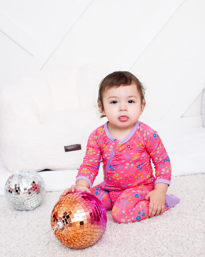 A toddler girl wearing a pink floral long-sleeve pajama set sits on a white carpeted floor, holding a shiny orange and pink disco ball. A smaller silver disco ball is on the left, with a white padded play couch and a pink fuzzy blanket in the background.