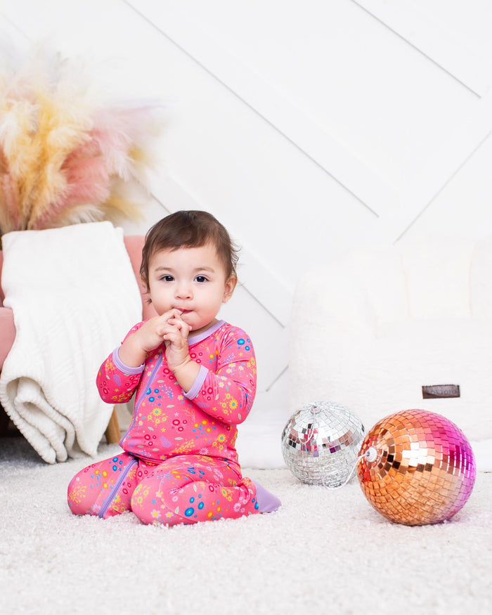 A toddler girl wearing a pink floral pajama set sits on a white carpet in a bright room, with a pink sofa, pampas grass, and two decorative disco balls nearby.