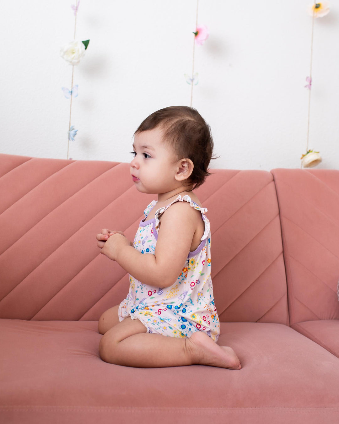 A toddler girl sitting on a dusty pink quilted sofa, wearing a sleeveless white romper with a colorful floral pattern. The background is a white wall decorated with hanging butterfly and flower garlands.