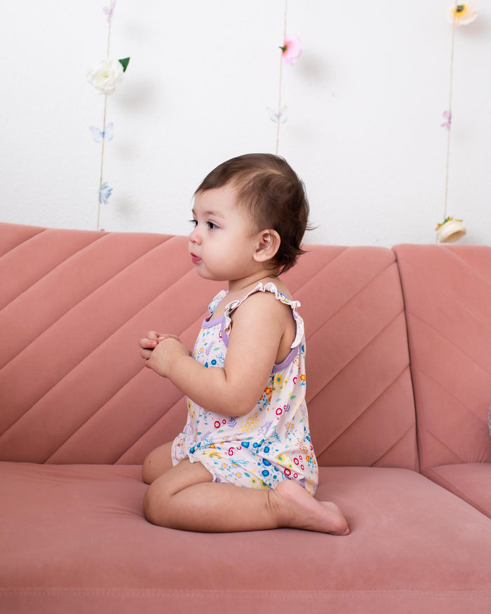 A toddler girl sitting on a dusty pink quilted sofa, wearing a sleeveless white romper with a colorful floral pattern. The background is a white wall decorated with hanging butterfly and flower garlands.