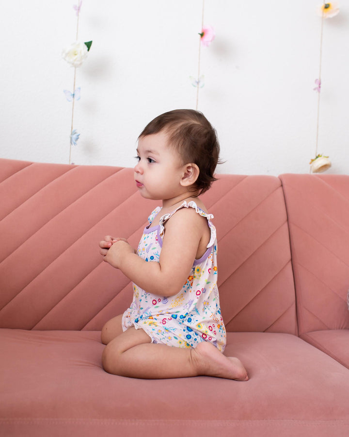 A toddler girl sitting on a dusty pink quilted sofa, wearing a sleeveless white romper with a colorful floral pattern. The background is a white wall decorated with hanging butterfly and flower garlands.