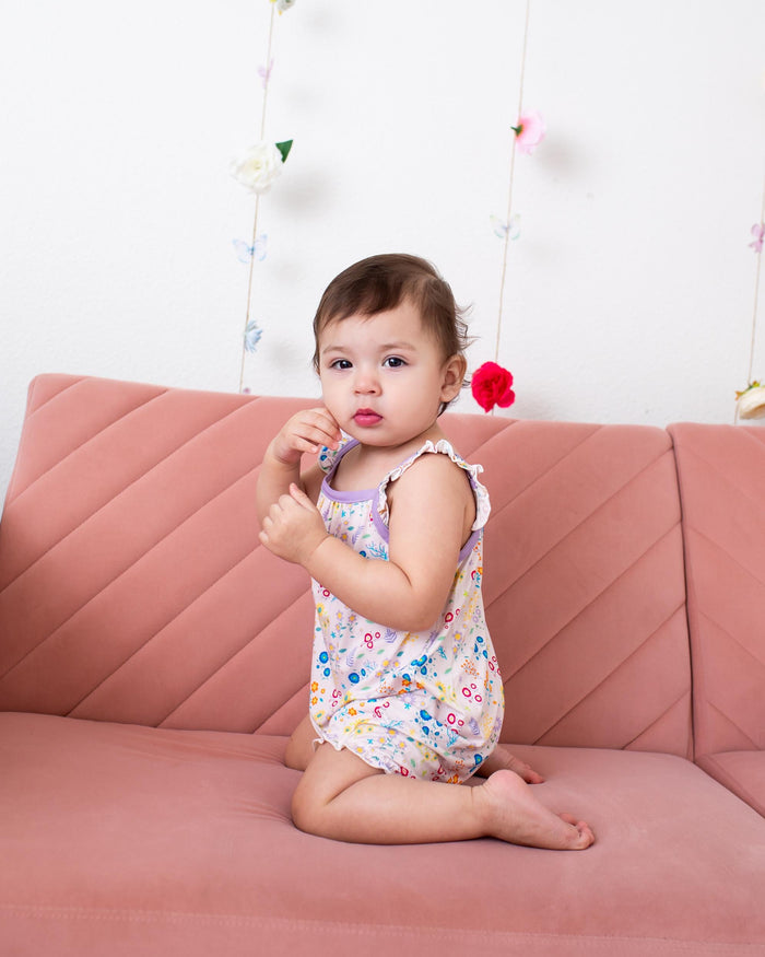 A baby girl sitting on a dusty pink sofa, wearing a sleeveless floral print romper. The background is a white wall decorated with hanging flower garlands.