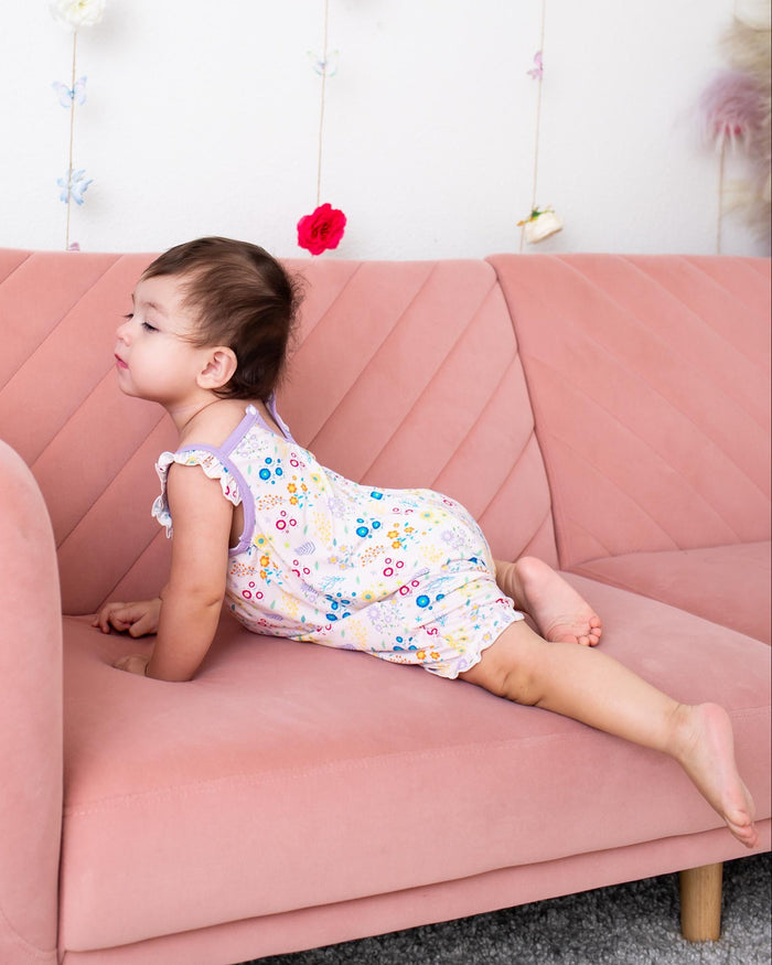 A young child wearing a pastel floral romper lies on a dusty pink velvet sofa in a brightly lit room with a white wall decorated with hanging flower garlands.