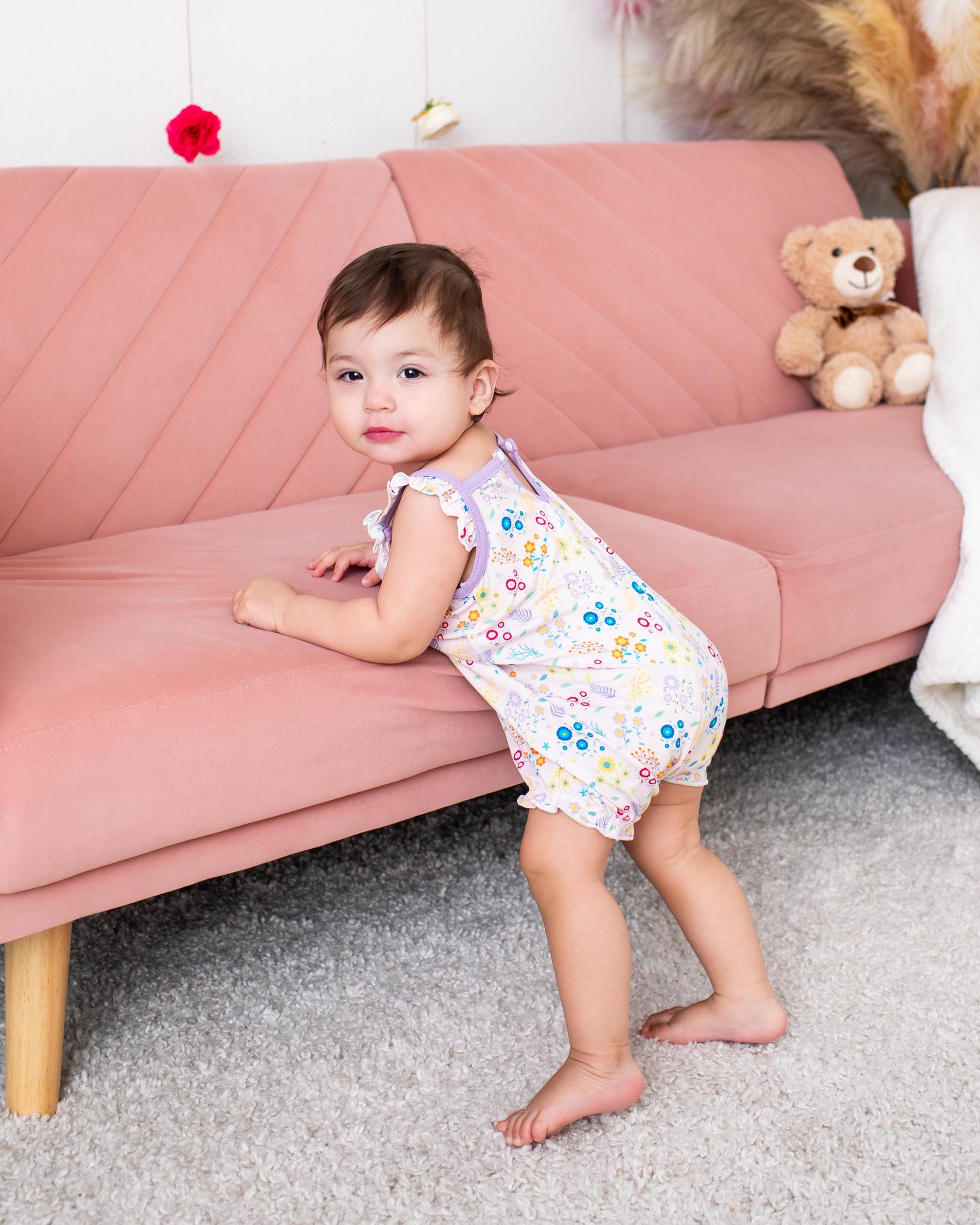 A toddler girl wearing a sleeveless white romper with a colorful floral print stands on a gray carpet in front of a dusty pink sofa. A teddy bear sits on the sofa, and pampas grass decor is visible in the background.