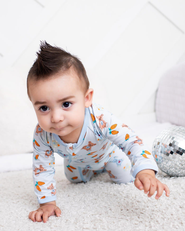 A baby crawling on a white carpet in a bright room, wearing a light blue long-sleeve pajama with a colorful cartoon animal print. A silver disco ball and a white pillow are visible in the background.