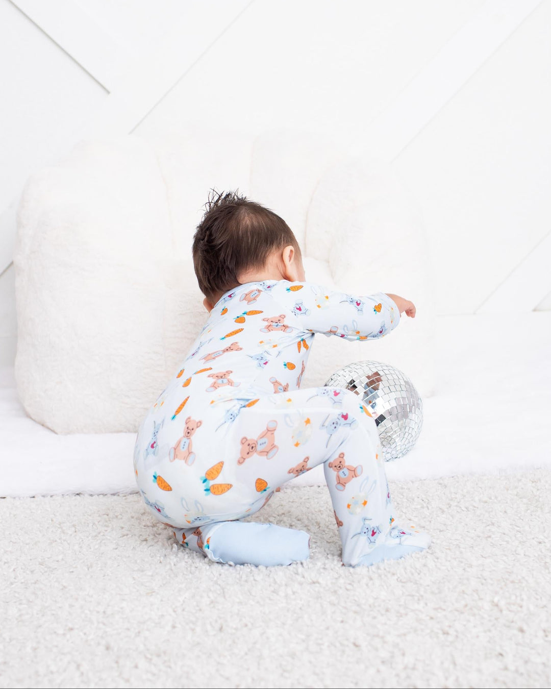 A baby wearing a light blue and white patterned footed pajama set crawls on a white carpet in a bright, modern nursery.