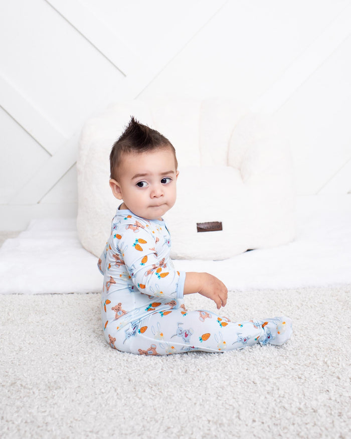 A baby sitting on a white carpet in front of a white padded chair, wearing a light blue footed onesie with a colorful leaf pattern.