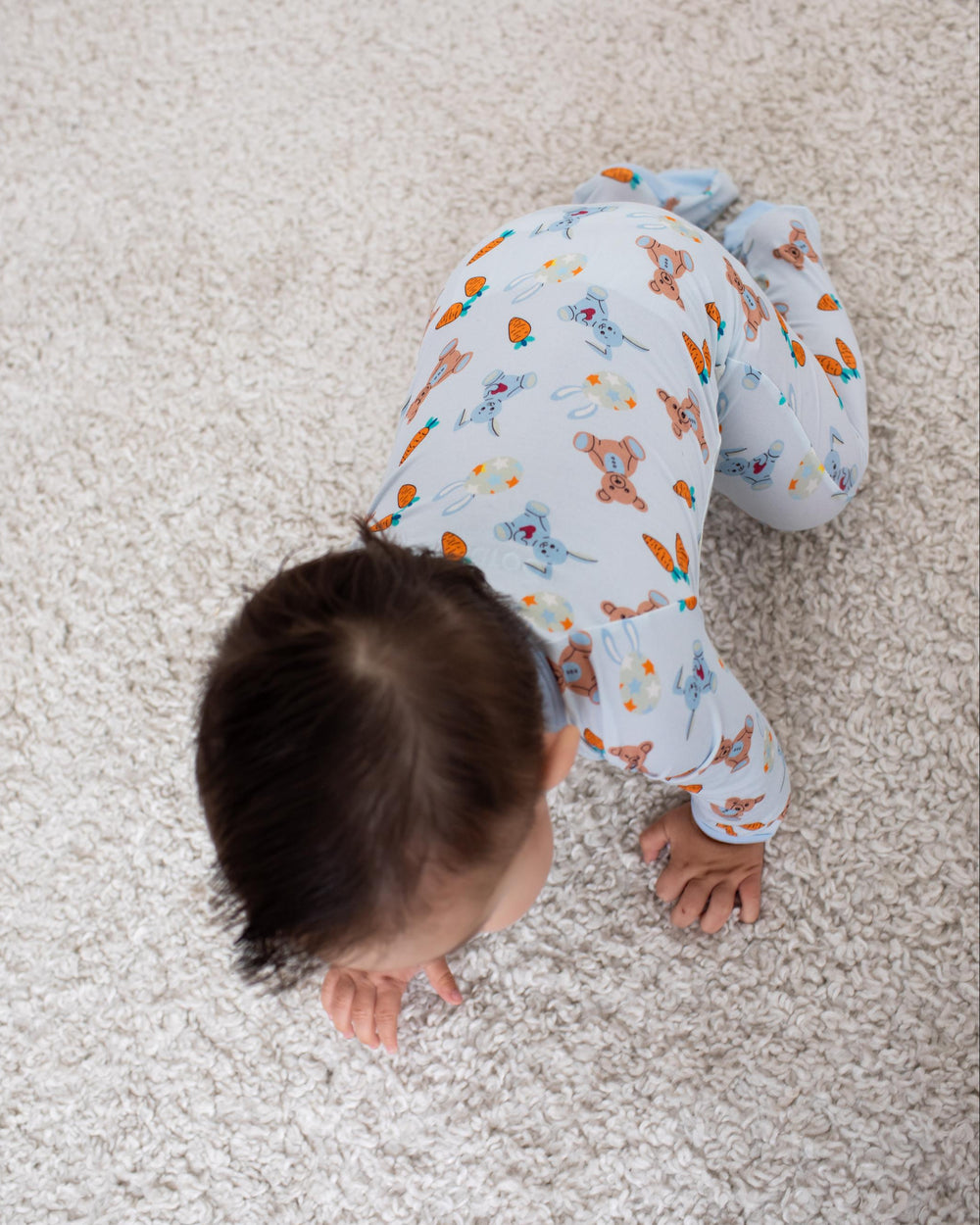 A baby crawling on a textured beige carpet, wearing a light blue long-sleeve pajama set with a colorful dinosaur pattern.
