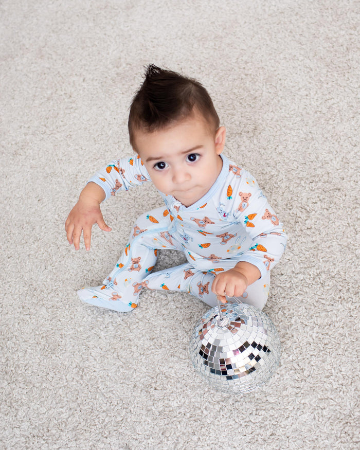A baby wearing a light blue and white patterned footed pajama sits on a light beige carpet, holding a small mirrored ball toy.