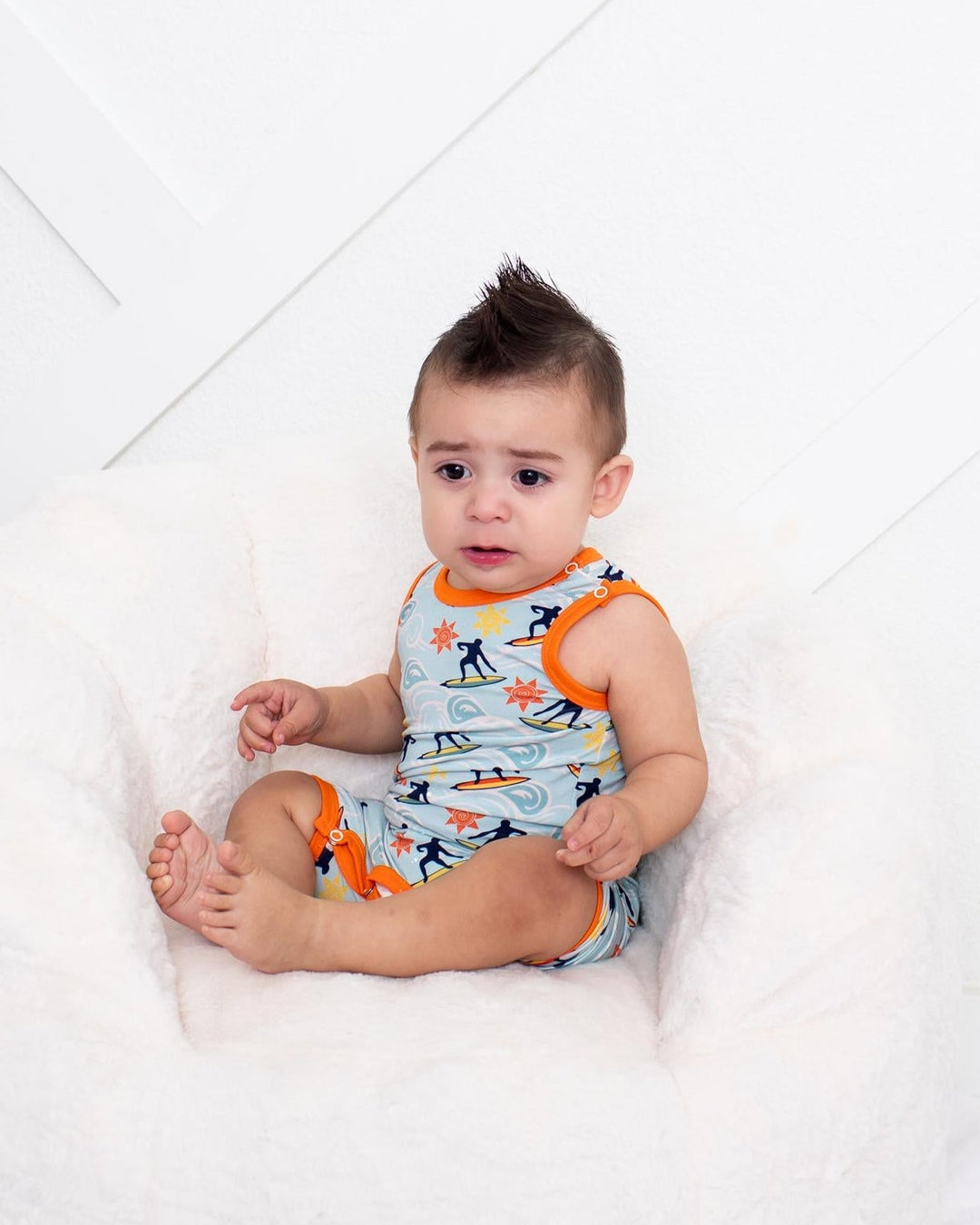 A baby sitting in a white plush bean bag chair, wearing a light blue and orange patterned romper, set against a white paneled wall.