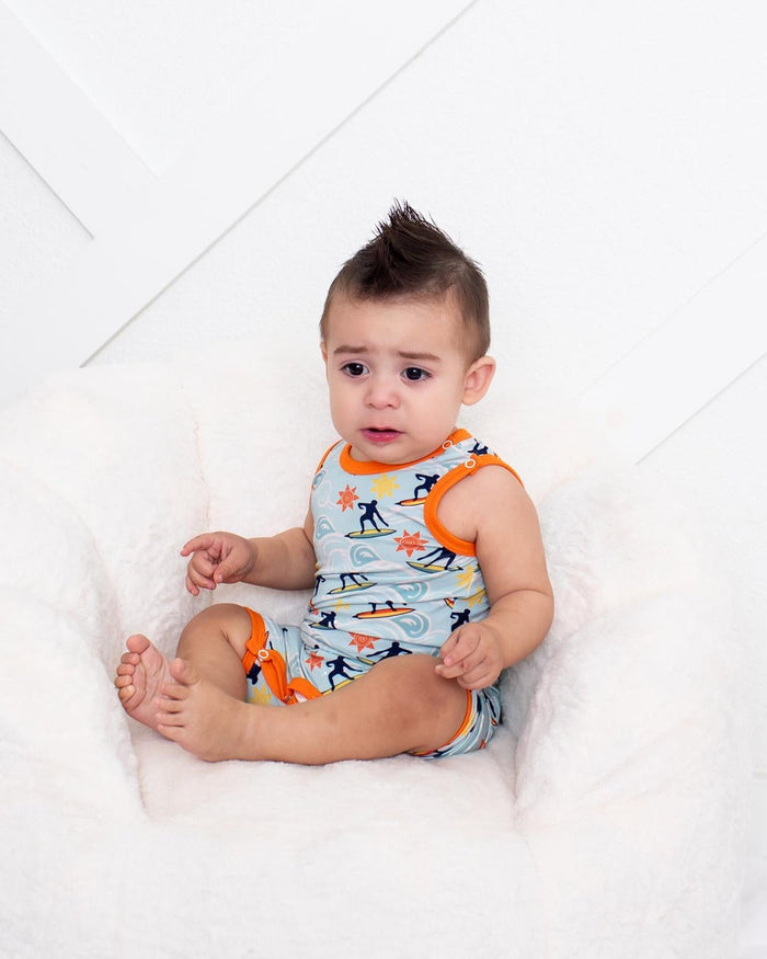 A baby sitting in a white plush bean bag chair, wearing a light blue and orange patterned romper, set against a white paneled wall.