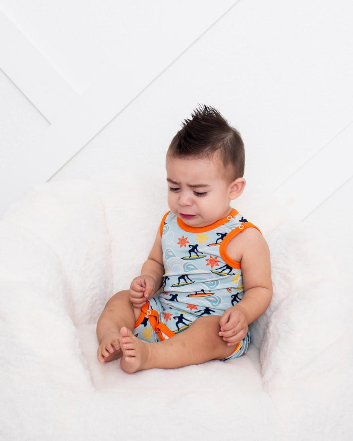 A baby sitting in a white plush beanbag chair, wearing a light blue and orange dinosaur print romper. A silver disco ball rests on the white floor in the foreground against a white paneled wall.