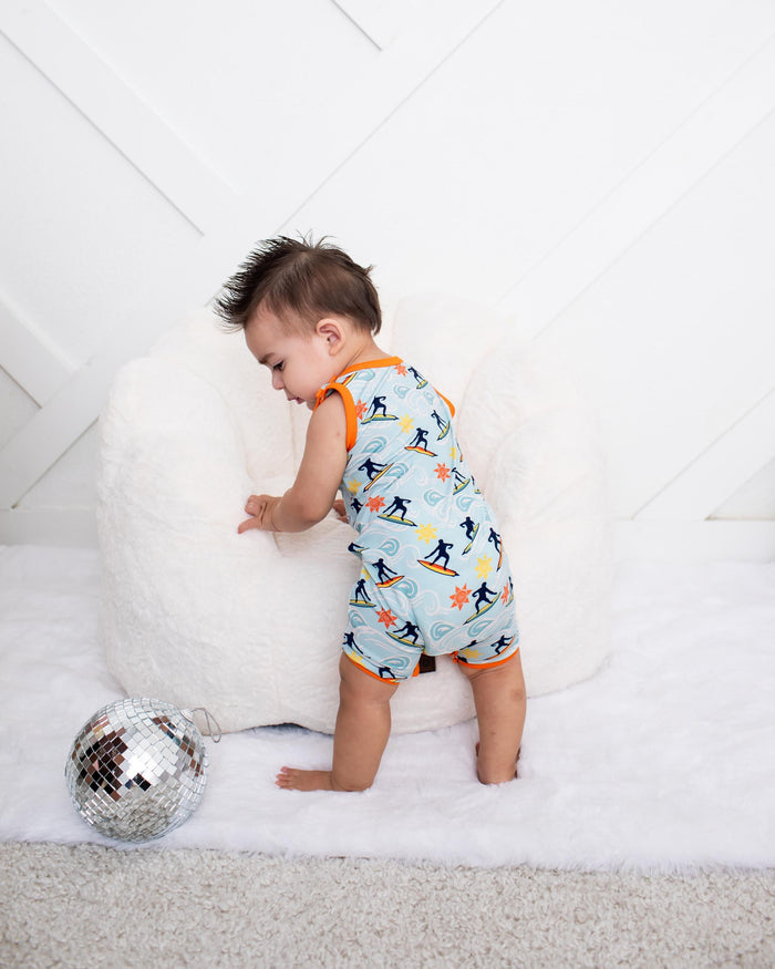 A toddler stands on a white rug next to a large white beanbag, wearing a light blue sleeveless romper with an orange collar and cuffs and a black-and-orange ski pattern. A silver disco ball sits on the rug in the foreground against a white paneled wall.