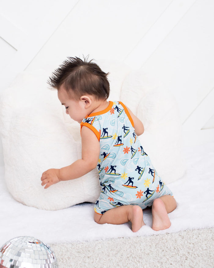 A baby wearing a light blue sleeveless romper with a black and orange penguin print sits on a white rug next to a white pillow in a bright room.