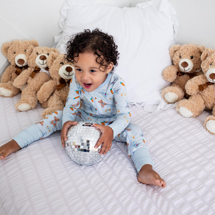 A toddler with curly dark hair sits on a white quilted bed, wearing light blue pajamas with a small animal print. The child is holding a small silver and white patterned ball, with several brown teddy bears lined up against a white pillow and wall behind them.
