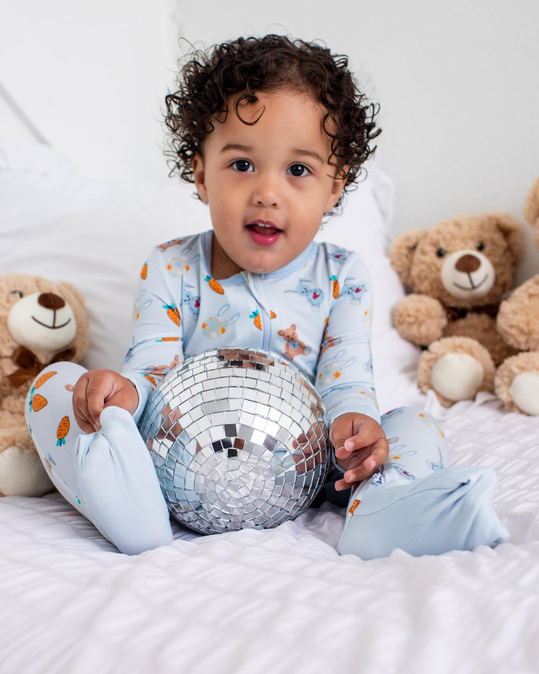 A toddler with curly dark hair sits on a white bed surrounded by teddy bears, wearing light blue pajamas with a small orange pattern and holding a silver disco ball.