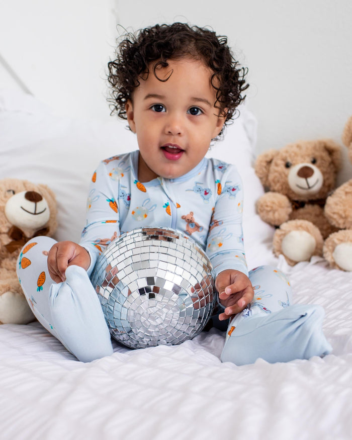 A toddler with curly dark hair sits on a white bed surrounded by teddy bears, wearing light blue pajamas with a small orange pattern and holding a silver disco ball.