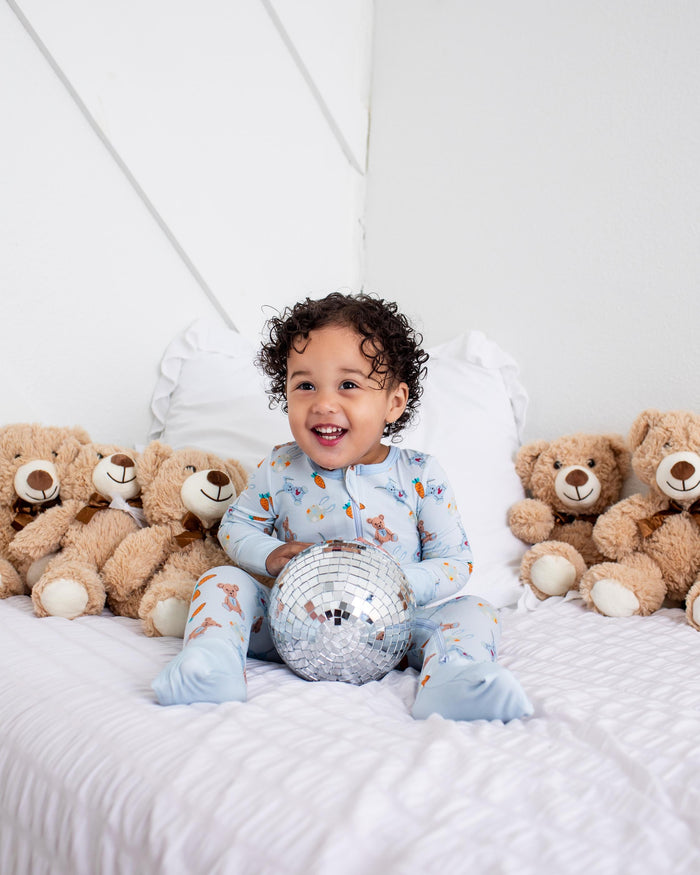 A toddler with curly dark hair sits smiling on a white bed, wearing light blue pajamas with a small animal print and holding a silver disco ball. Brown teddy bears line both sides of the bed against a white wall.