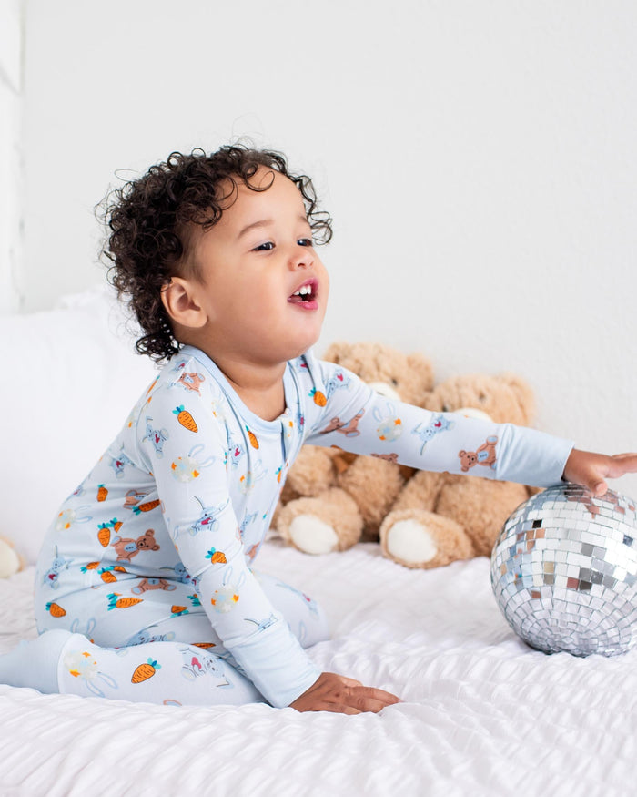 A toddler with curly dark hair sits on a white bed wearing light blue pajamas with a colorful animal print. The child is reaching toward a silver disco ball, with plush teddy bears lined up against a white headboard in the background.