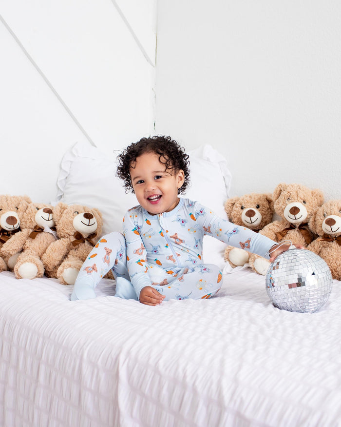 A smiling toddler wearing light blue patterned pajamas sits on a white bed surrounded by several teddy bears, with a small gray plush toy in front of them.