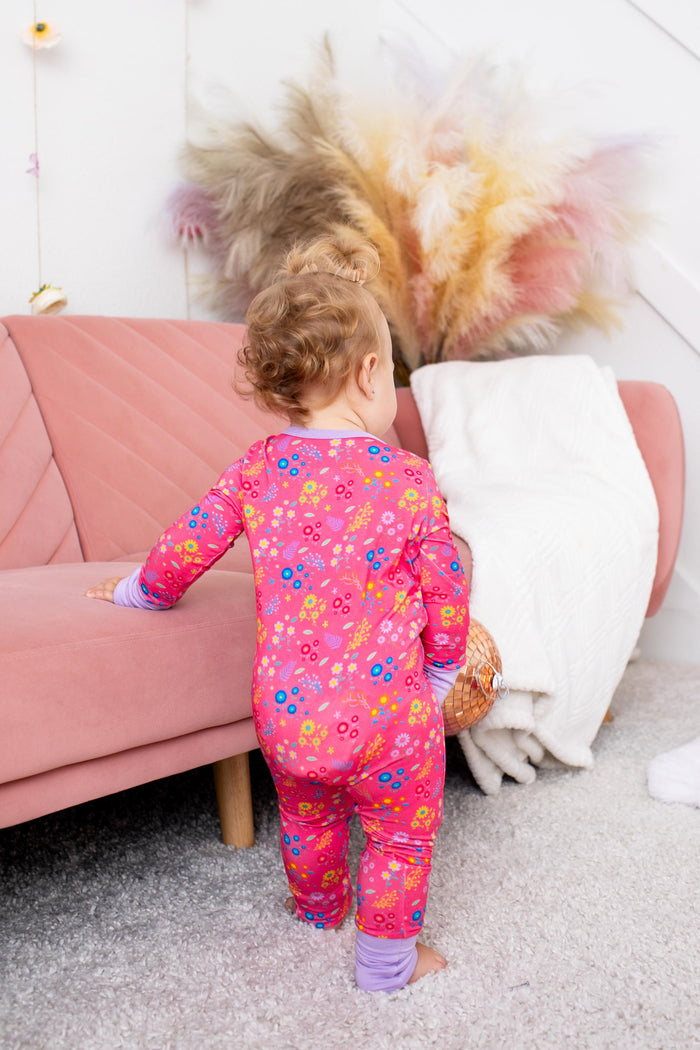 A toddler seen from behind wearing a bright pink long-sleeve pajama set with a colorful floral print, standing in a cozy room next to a pink sofa.