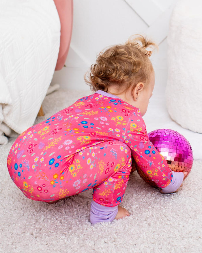 A toddler in a pink floral long-sleeve pajama set crouches on a light carpet, holding a shiny pink disco ball toy in a cozy room with white beanbag and soft furnishings.