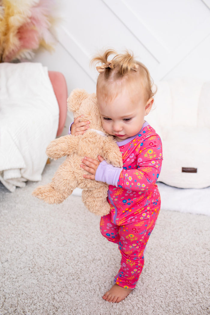 A toddler standing barefoot on a light carpet and holding a beige teddy bear, wearing a bright pink one-piece pajama with colorful floral patterns and a light purple cuff.
