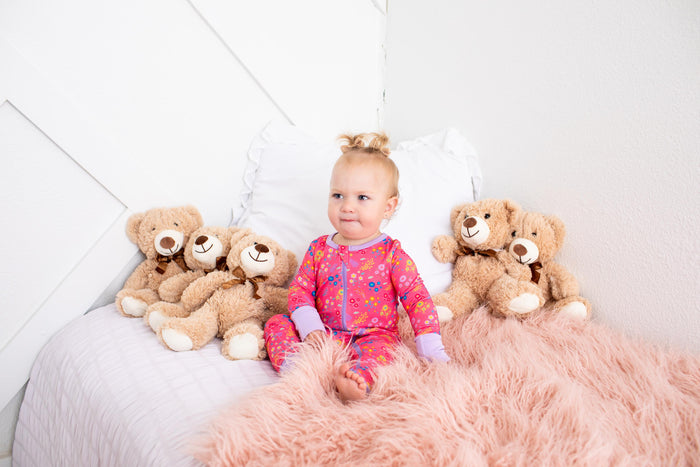 A baby girl wearing a pink floral pajama set sits on a bed with white pillows and a fluffy pink blanket, surrounded by four brown teddy bears.