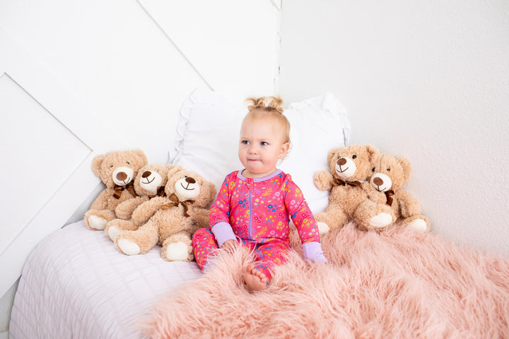 A baby girl wearing a pink floral pajama set sits on a bed with white pillows and a fluffy pink blanket, surrounded by four brown teddy bears.